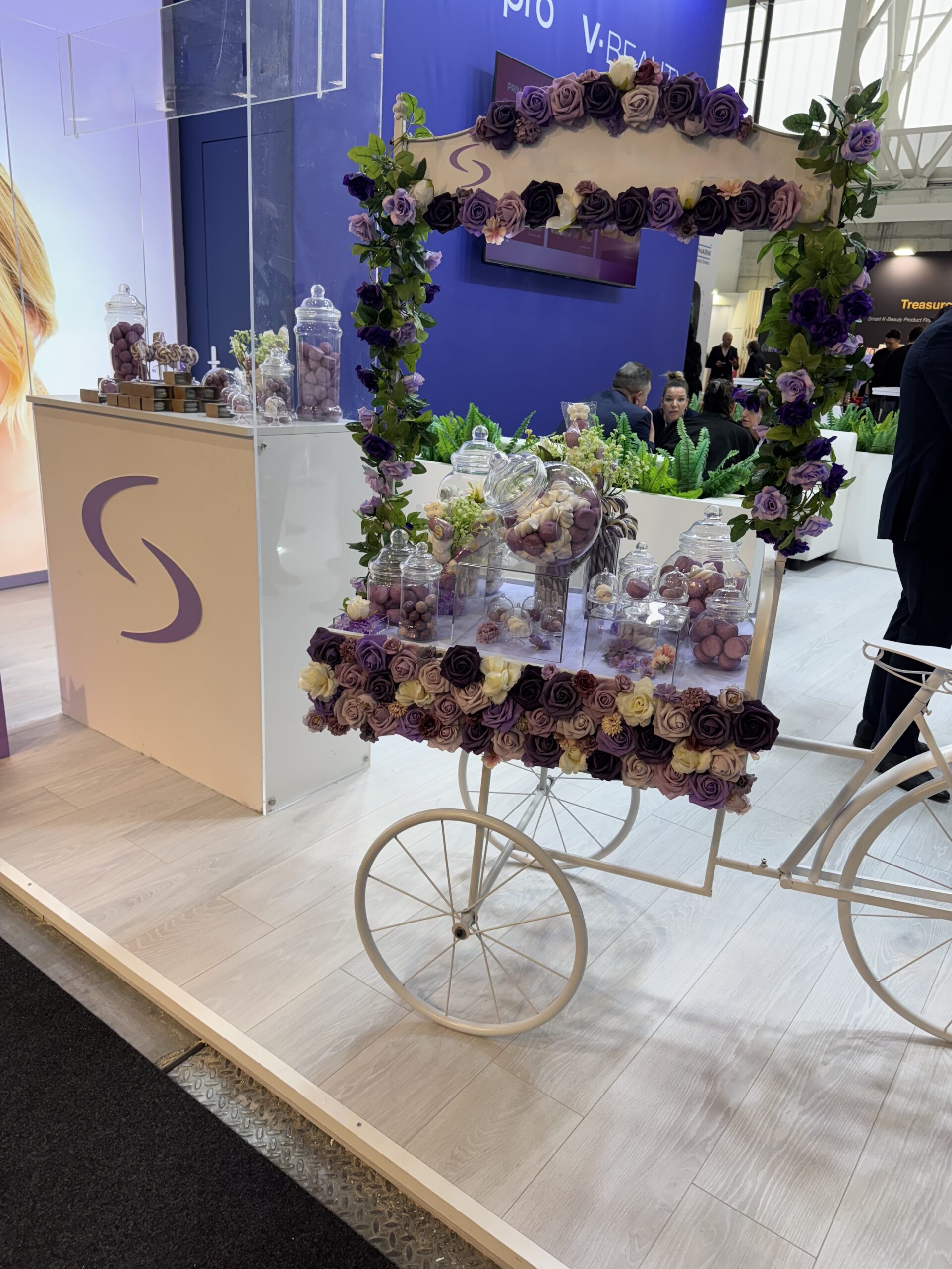 Mit Blumen geschmückter Präsentationswagen mit Glasgefäßen und violetten Blüten auf der Cosmoprof Bologna 2025, eleganter Messestand mit kosmetischem Fokus.